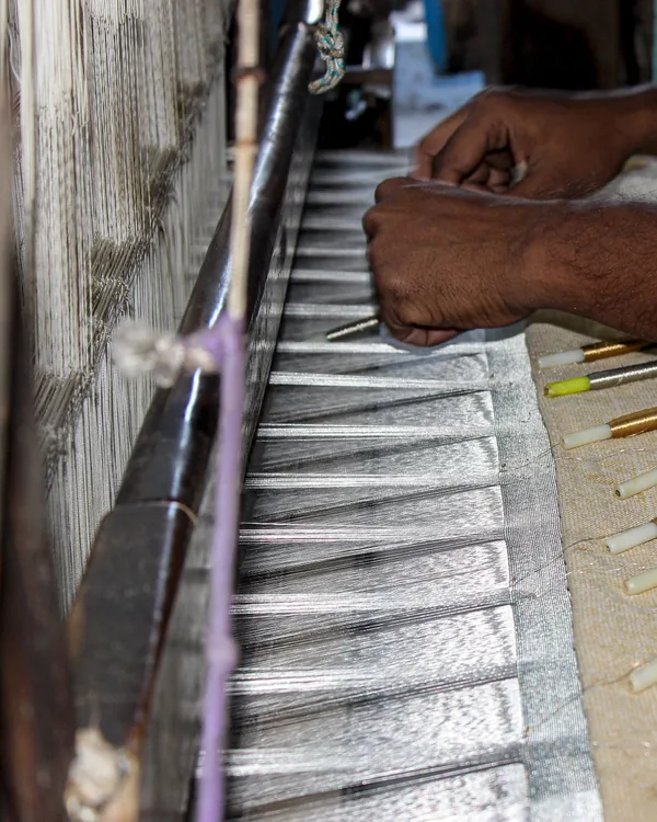 A weaver's hands at the reed of a loom, pressing the last pick into the cloth.